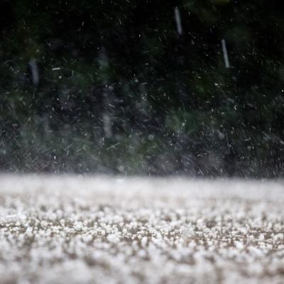 Close-up of hailstones falling and accumulating on the ground, with blurred greenery in the background.