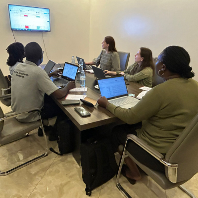 Five people sitting around a conference table with laptops, viewing a large screen displaying a spreadsheet in a meeting room.