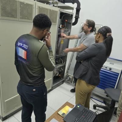 Three people stand in front of open server equipment in a lab, discussing the components; a laptop and documents are on a nearby table.