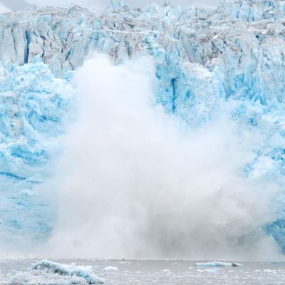 Large chunk of glacier ice collapsing into the water, creating a splash and mist at the base, with blue and white ice formations in the background.