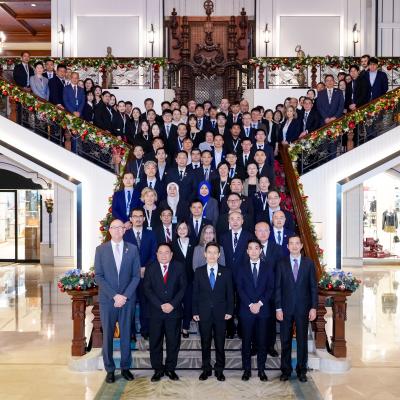 A large group of formally dressed people pose for a group photo on a grand staircase decorated with festive garlands in an ornate building lobby.