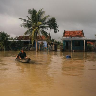 A person paddles a small boat through floodwaters in front of partially submerged homes and palm trees under a cloudy sky.