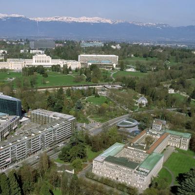 Aerial view of several large buildings surrounded by green lawns and trees, with mountains and snow-capped peaks visible in the background.