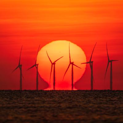 Wind turbines are silhouetted against a large setting sun over the ocean, with the sky glowing in shades of orange and red.