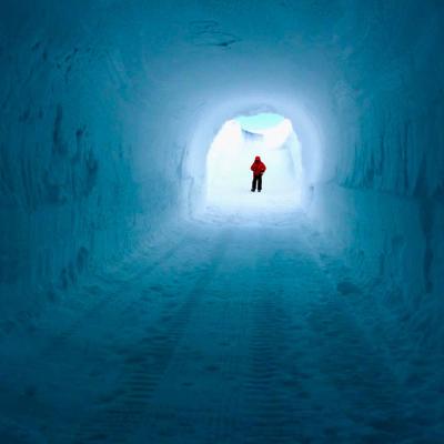A person wearing a red jacket stands at the end of a tunnel made of snow and ice, illuminated by natural light.