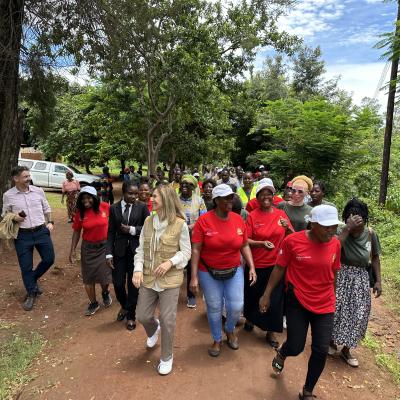 A group of people, including women in red shirts and white caps, walk together outside on a tree-lined path, with others and a white van visible in the background.