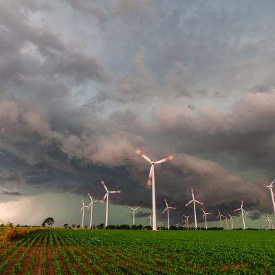 Wind turbines in a field with storm clouds overhead, green crops in the foreground, and dramatic sky in the background.