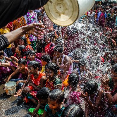 A group of children sit closely together as water is poured over them from a bucket, surrounded by a crowd of onlookers in colorful clothing.