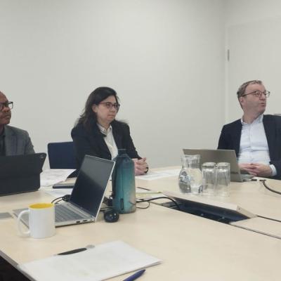 Three people sit at a conference table with laptops, notebooks, a water bottle, and glasses, appearing to attend a meeting in a modern office setting.