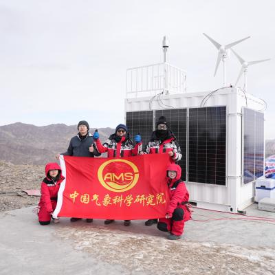 Four people in red jackets hold a red flag with Chinese text and "AMS" logo in front of a solar- and wind-powered research station on a rocky mountain.