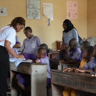 A teacher assists students in a classroom as another adult observes; students are seated at wooden desks wearing purple uniforms. Educational posters are on the walls.