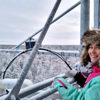 A person in a fur-lined hooded winter coat stands on a metal structure overlooking a snowy forest, smiling at the camera.