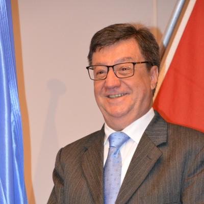 A man in a suit and glasses smiles while standing in front of blue and red flags.