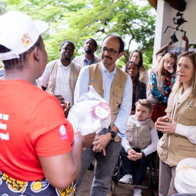 A group of people, some wearing vests, listen attentively to a person in a red shirt holding supplies during an outdoor gathering.