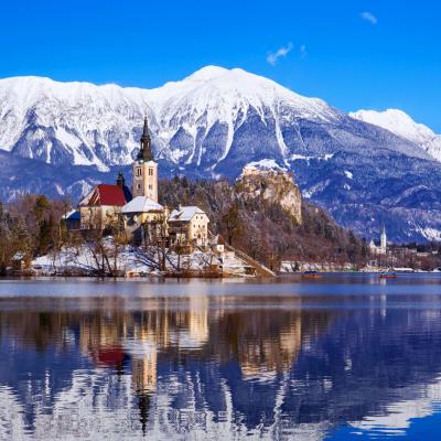 A church and castle stand on Lake Bled’s island, surrounded by snow-capped mountains and reflected in the calm water under a clear blue sky.