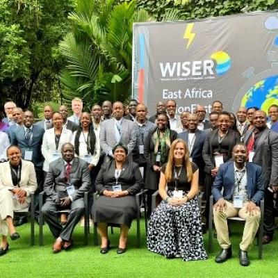 A large group of people pose for a photo outdoors in front of a sign that reads "WISER East Africa," surrounded by greenery.