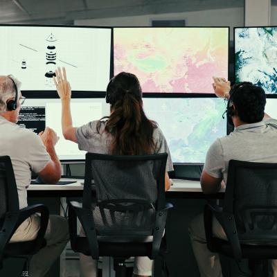 Three people wearing headsets sit at desks, facing large monitors displaying maps, satellite images, and data visualizations in a modern control room.
