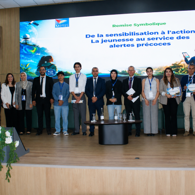 A group of people in business attire stand on stage in front of a screen displaying a French presentation about youth involvement in early warning systems.
