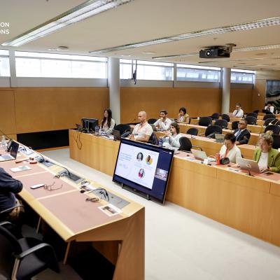 People attend a conference in a modern meeting room, with speakers at a front panel and a large screen displaying a presentation; GEO Group on Earth Observations logo is visible.