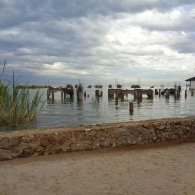 Sandy path with a stone wall, water with old wooden pilings, reeds on the left, and a small white building on the right under a cloudy sky.