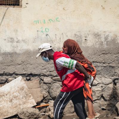 A person in a red vest and face mask carries another person on their back along a rough, debris-strewn street next to a worn wall.