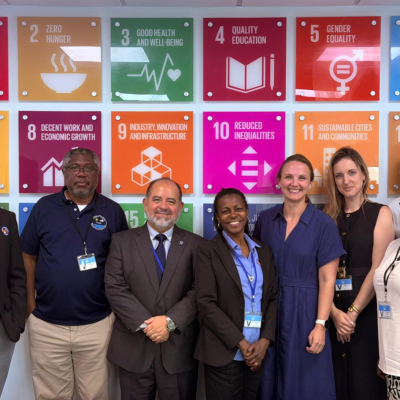 Eight people stand smiling in front of a colorful wall displaying icons and goals related to the United Nations Sustainable Development Goals.