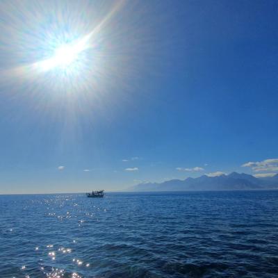 The sun shines brightly over a calm blue sea with a boat in the distance and mountains visible on the horizon under a clear sky.