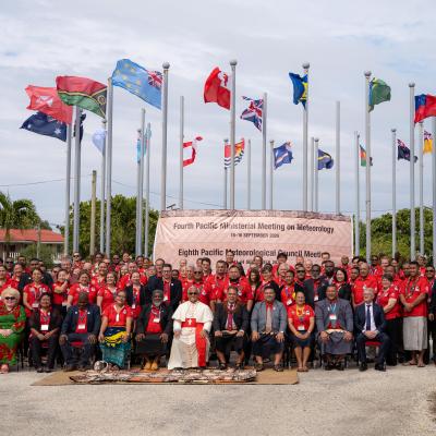 A large group of people pose for a photo in front of international flags and a banner for the Pacific Meteorological meeting, with palm trees and a clear sky in the background.