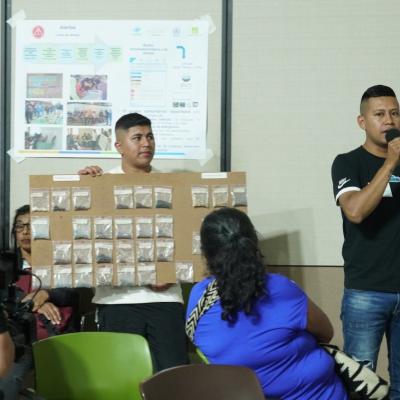 A man speaks into a microphone while another holds a display board with many small bags. Several people are seated, and posters with images and charts are on the wall behind them.