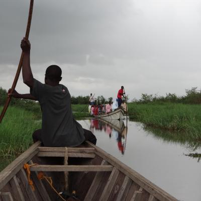 A person steers a wooden boat with a long pole through a grassy channel, following another boat filled with people.