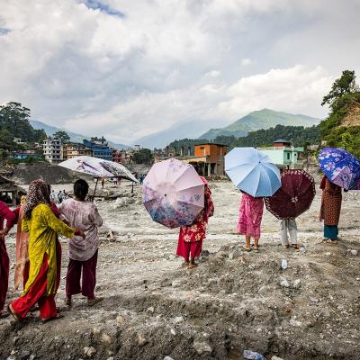 People holding umbrellas stand on a rocky, debris-filled landscape overlooking a town near mountains.