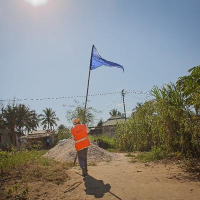 A person in an orange vest carries a tall pole with a blue flag on a dirt path, with trees and houses in the background under a clear sky.