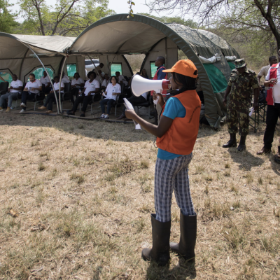 A person in an orange vest speaks through a megaphone to a group of people seated under a large canvas tent in a field.