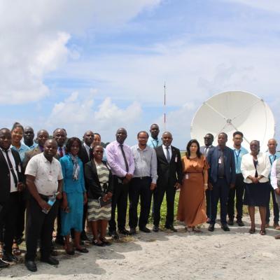 A group of people poses outdoors in front of satellite dishes on a grassy area, dressed in business and casual attire under a partly cloudy sky.