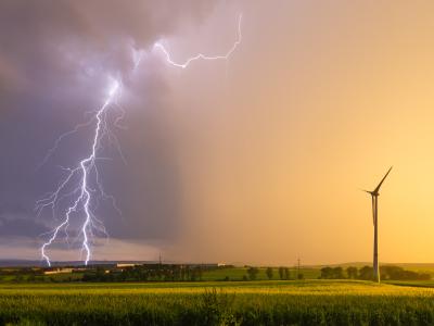 A lightning bolt strikes in the distance over fields at sunset, with a single wind turbine standing in the foreground.