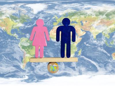 Pink female and blue male symbols stand balanced on a wooden plank over a globe, with a world map background, representing gender equality worldwide.