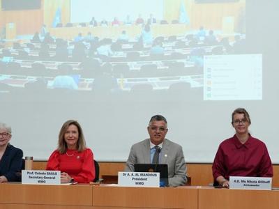 Six people sit at a conference table with nameplates, facing the camera, with a large screen showing an audience and meeting behind them.