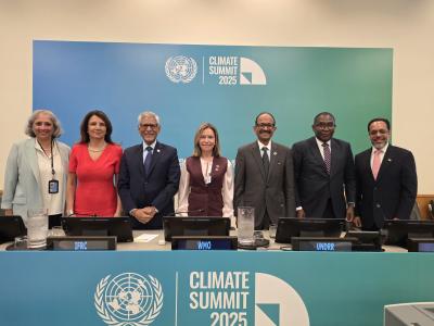 Seven delegates stand behind a table at the Climate Summit 2025, with UN and organization logos displayed on the wall and table in a conference room setting.