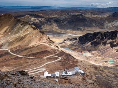 A remote mountainous landscape with winding dirt roads, a cluster of white buildings in the foreground, and rugged, rocky hills and small turquoise lakes in the background.