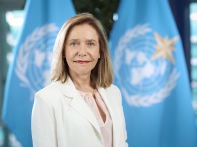 A woman in a white blazer stands in front of two blue flags featuring the United Nations emblem.
