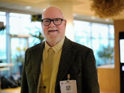 Bald man with glasses, beard, and mustache wears a suit, yellow shirt, and tie, standing indoors with a conference name badge clipped to his jacket.