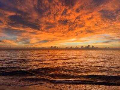 Orange and gold clouds fill the sky over a calm ocean at sunset, reflecting warm colors on the water and gentle waves near the shore.