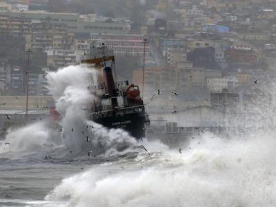 A ship being struck by large waves in a harbor, with a hillside city in the background.