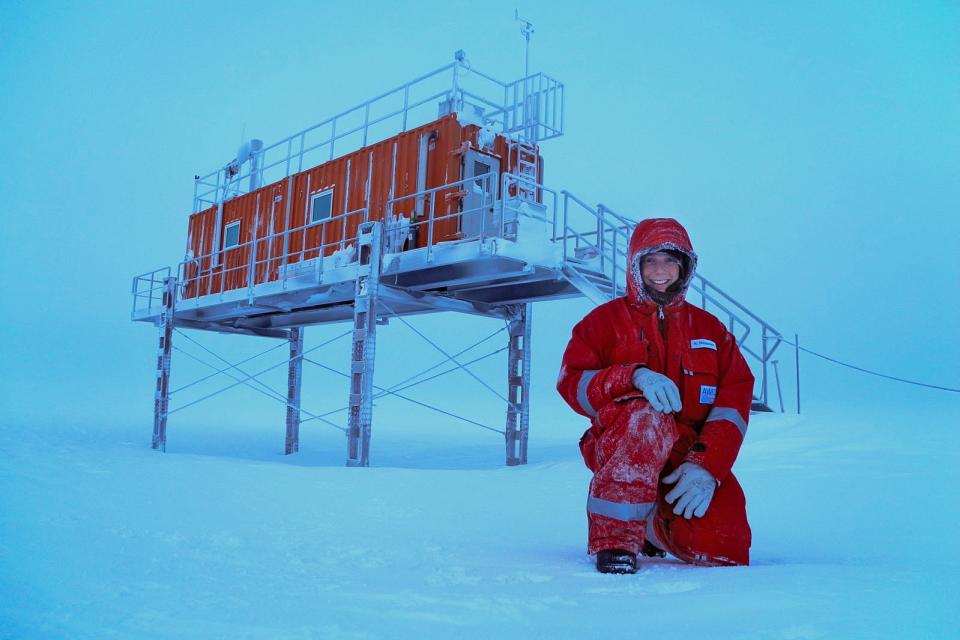 A person in a red winter suit kneels in snowy conditions in front of an elevated orange structure on metal stilts.