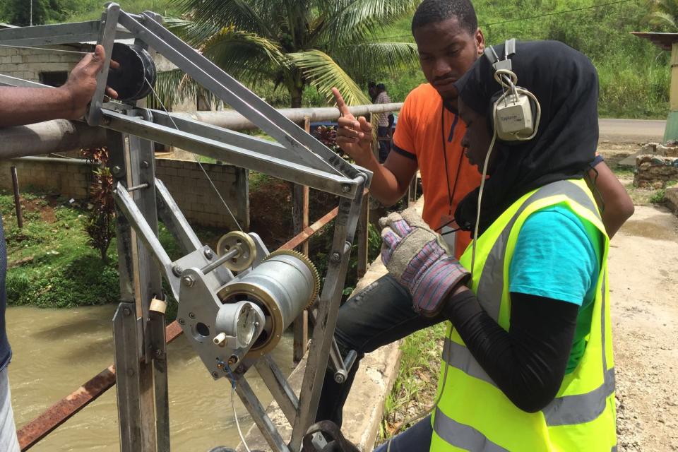 Two people operate a metal winch mechanism beside a river; one wears a safety vest, gloves, and earmuffs, while the other provides instructions.