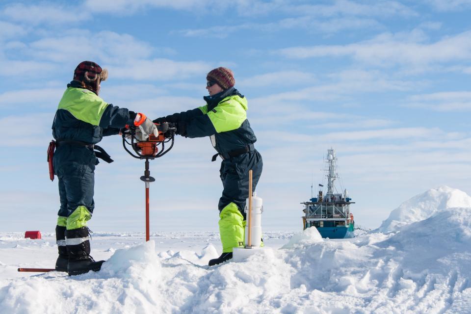 Two people in winter gear use a drilling tool on ice, with scientific equipment nearby and a research vessel in the background under a partly cloudy sky.