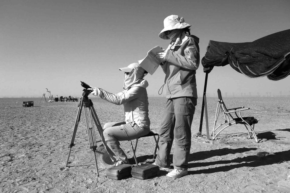 Two people on a sandy field; one is seated using binoculars and the other stands holding a clipboard. Both wear hats and sun-protective clothing. A tent and chair are nearby.