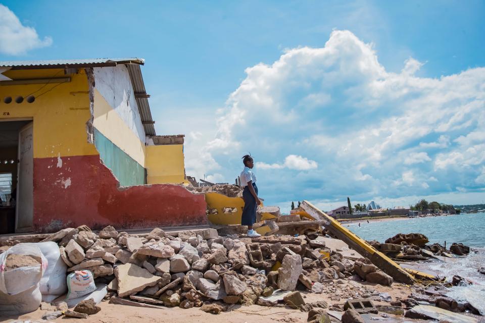 A person stands near a partially collapsed building with rubble scattered around, next to a body of water under a partly cloudy sky.