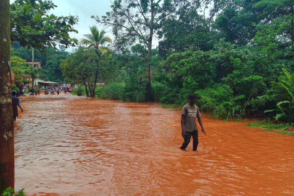 A person wades through a flooded street surrounded by dense green vegetation and trees, with more people visible in the background.