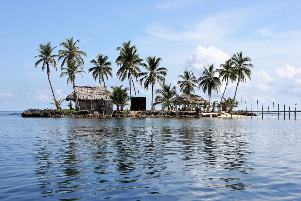 Small tropical island with palm trees, thatched huts, and a wooden dock, surrounded by calm blue water under a partly cloudy sky.